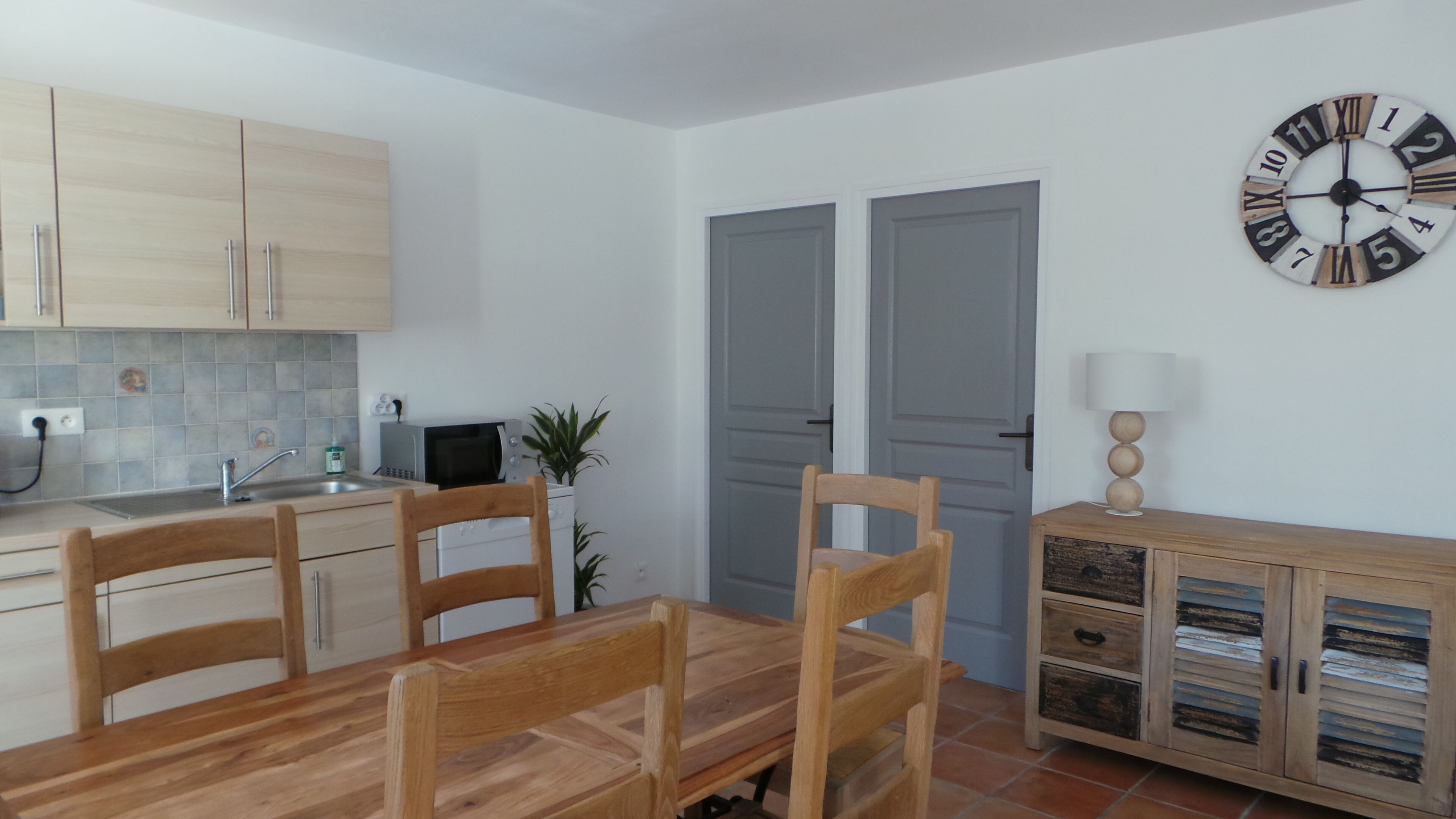 A compact kitchen with wooden cabinets, a dining table with chairs, a clock, and a cabinet with drawers.
