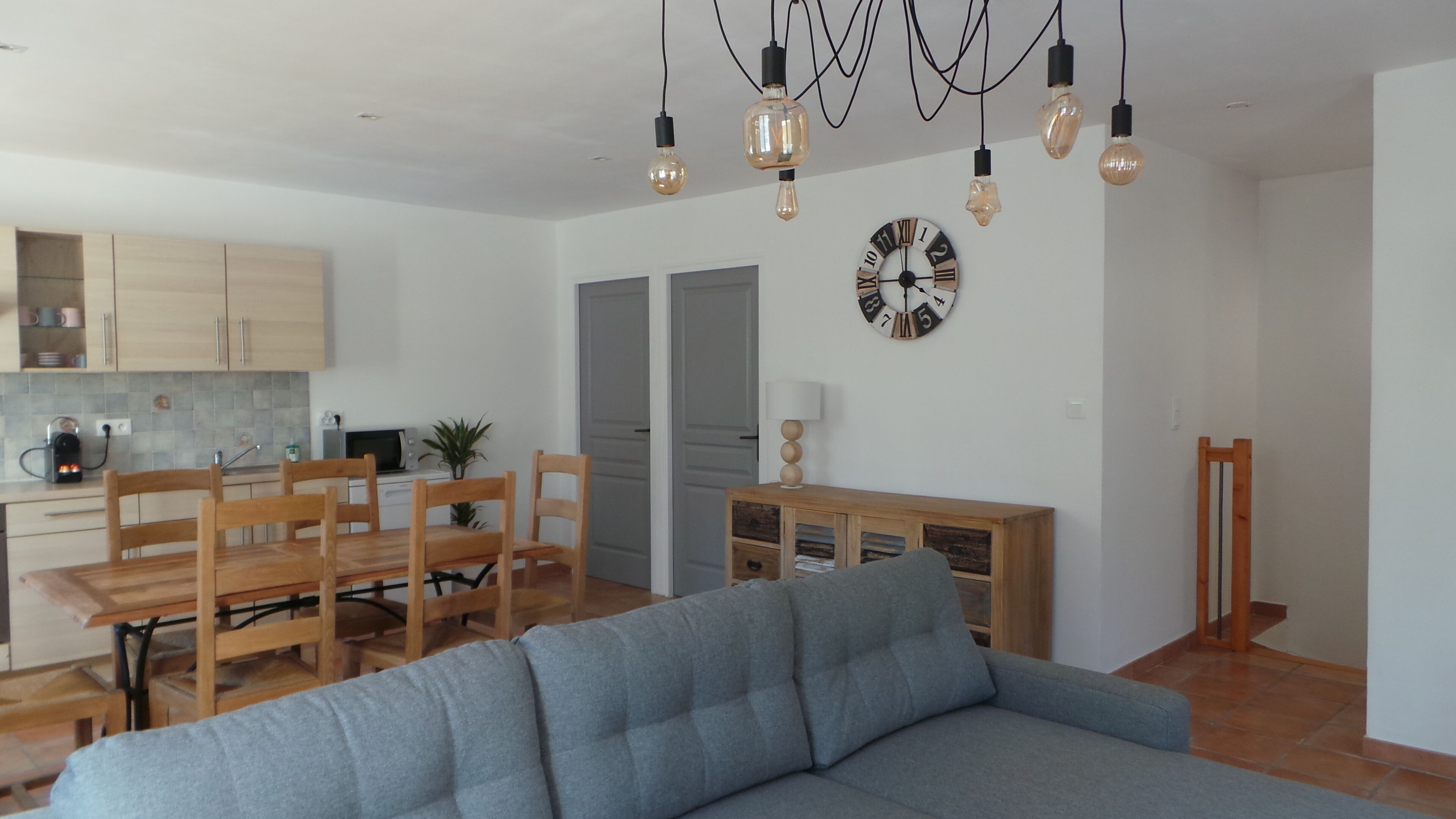 Een moderne woonkamer met een grijze bank, een houten eettafel en stoelen. Er is een keuken met kasten en een magnetron. Een klok en een lamp hangen aan de muur.