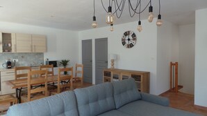 Una sala moderna con un sofá gris, una mesa de comedor de madera y sillas. Hay una zona de cocina con armarios y un microondas. Un reloj y una lámpara están en la pared.