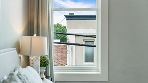 A bedroom with a window, a bed, a bedside table with a lamp, and a view of a building outside. - Downtown Cartersville 1BR Loft 1 (Cartersville)