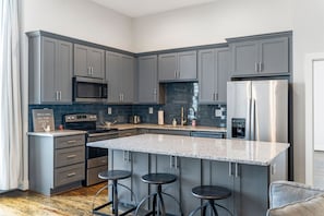 A modern kitchen with grey cabinets, stainless steel appliances, a granite countertop, and three stools.