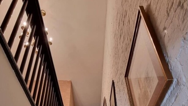 Eine Treppe mit Holzdielen und einem schwarzen Geländer, die zu einem Zwischenpodest mit einer weißen Backsteinwand und gerahmten Bildern führt.