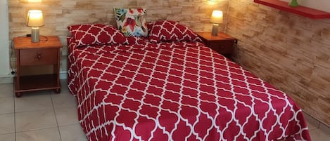 A bedroom with a beach-themed mural, a red patterned bedspread, two bedside lamps, a wooden headboard, and a red shelf with a green plant.