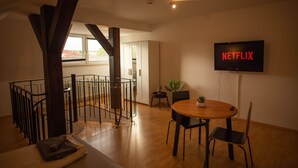 Un salon moderne avec une table à manger en bois, des chaises noires, un téléviseur affichant le logo « NETFLIX » et un escalier doté d’une rampe en métal.