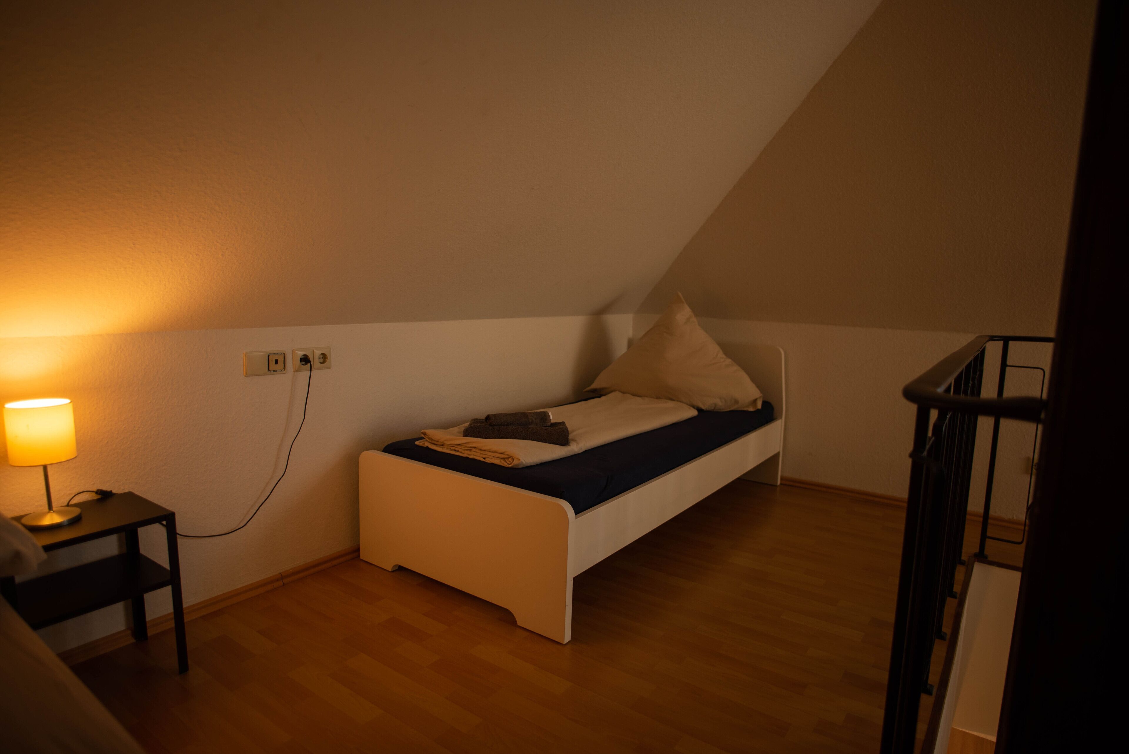 Une petite chambre avec un lit simple, un plancher en bois, une table de chevet avec une lampe et une prise électrique au mur.