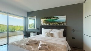 Ein modernes Schlafzimmer mit einem großen Bett, einem Schreibtisch und Blick auf einen Strand durch ein großes Fenster.