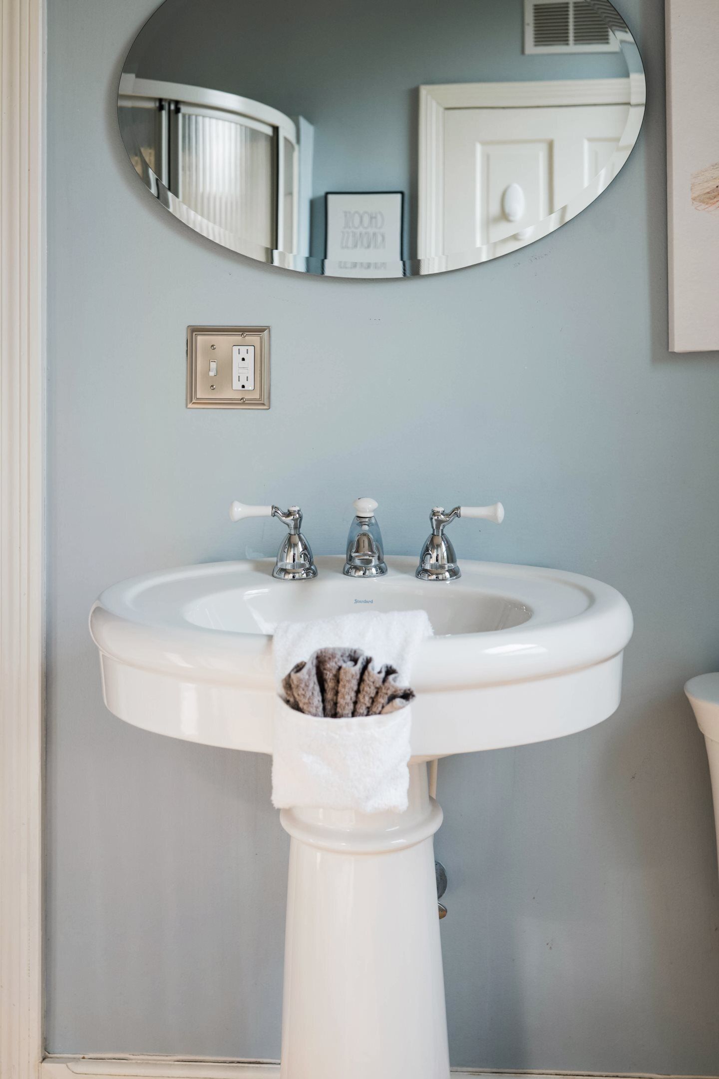 A bathroom with a white pedestal sink, a mirror above it, and a framed picture on the wall.