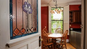 A dining area with a round table and wooden chairs, a stained glass window, and a view of greenery outside. - Historic Downtown Manistee Apartment (Manistee)