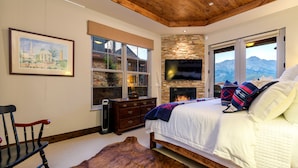 Une chambre avec une cheminée en pierre, un lit avec une literie blanche, une tête de lit en bois et une grande fenêtre donnant sur la montagne.