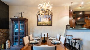 Un coin repas avec une table en bois dressée pour six personnes, entourée de chaises. Un grand tableau représentant une scène pastorale est accroché au-dessus de la table.