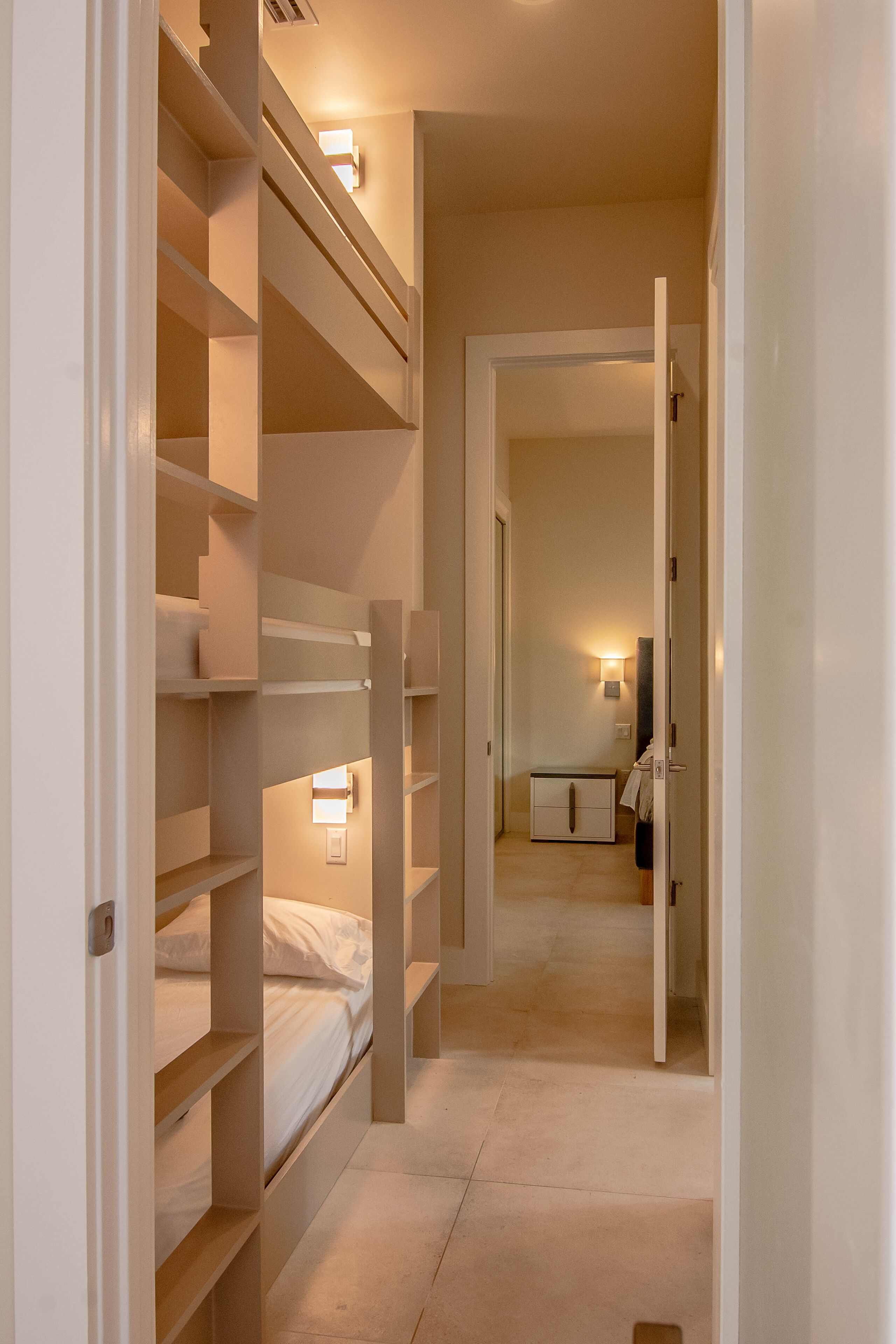 A narrow hallway with bunk beds on one side and a door leading to a room with a bed and a nightstand.