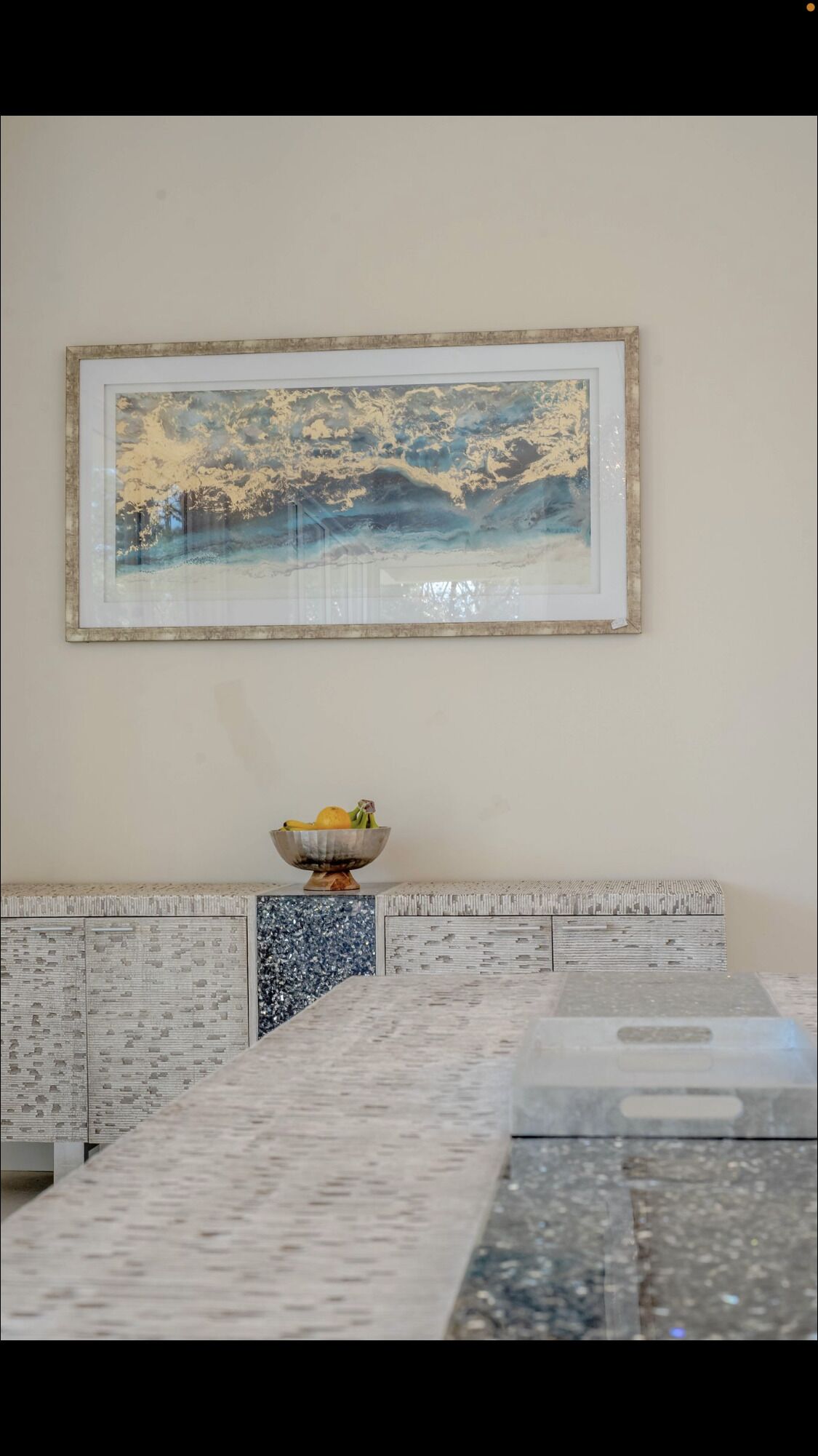 A kitchen with a marble countertop, a framed painting above the sink, and a bowl of fruit on the counter.