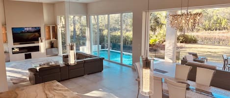 A modern living room with a large TV, a pool, and a dining area with outdoor seating.