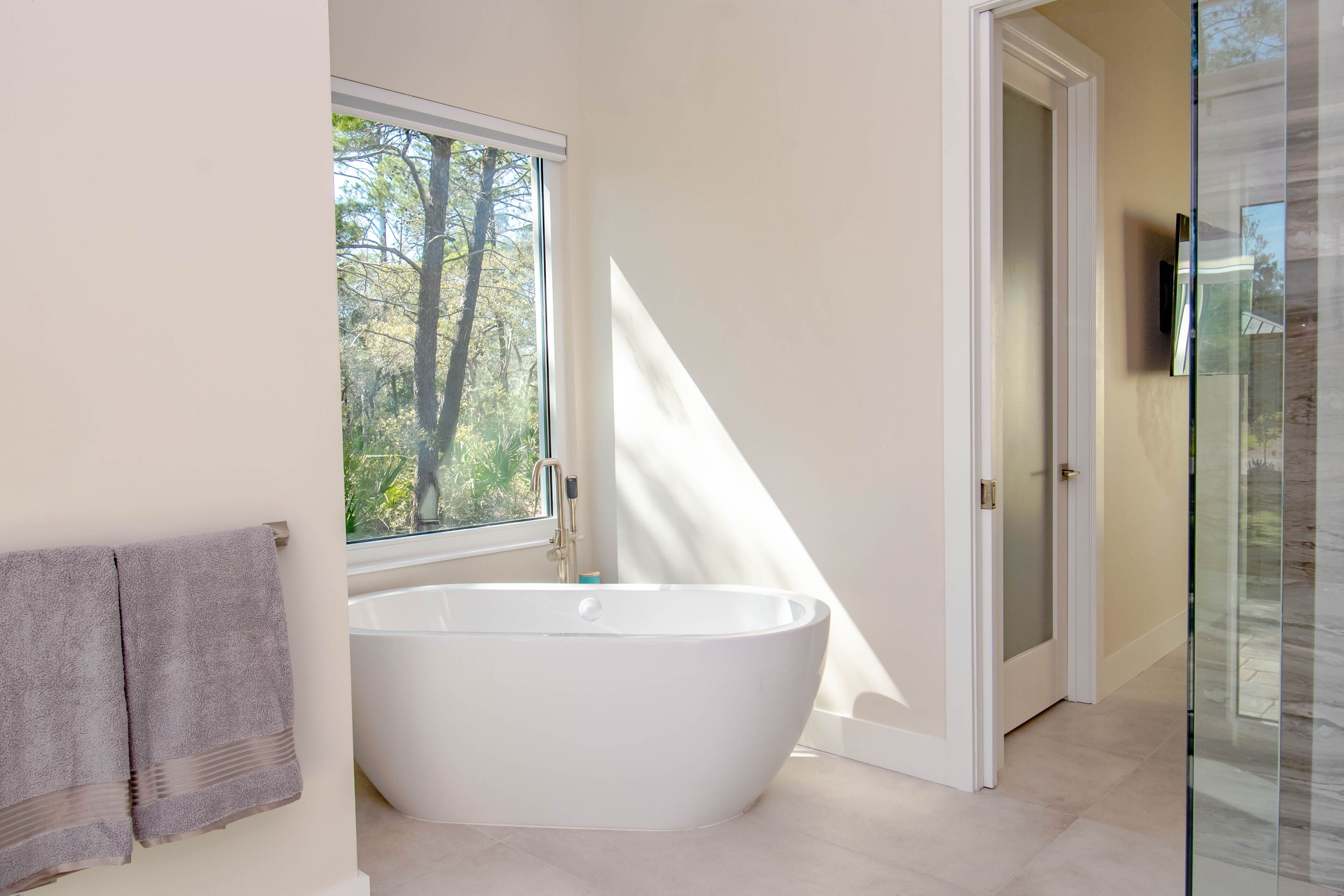 A modern bathroom with a freestanding bathtub, a window with a view of trees, and a glass shower enclosure.