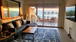 Un salon avec un canapé, une table basse et un tapis à motifs. Il y a un balcon avec vue sur l’océan et les palmiers.