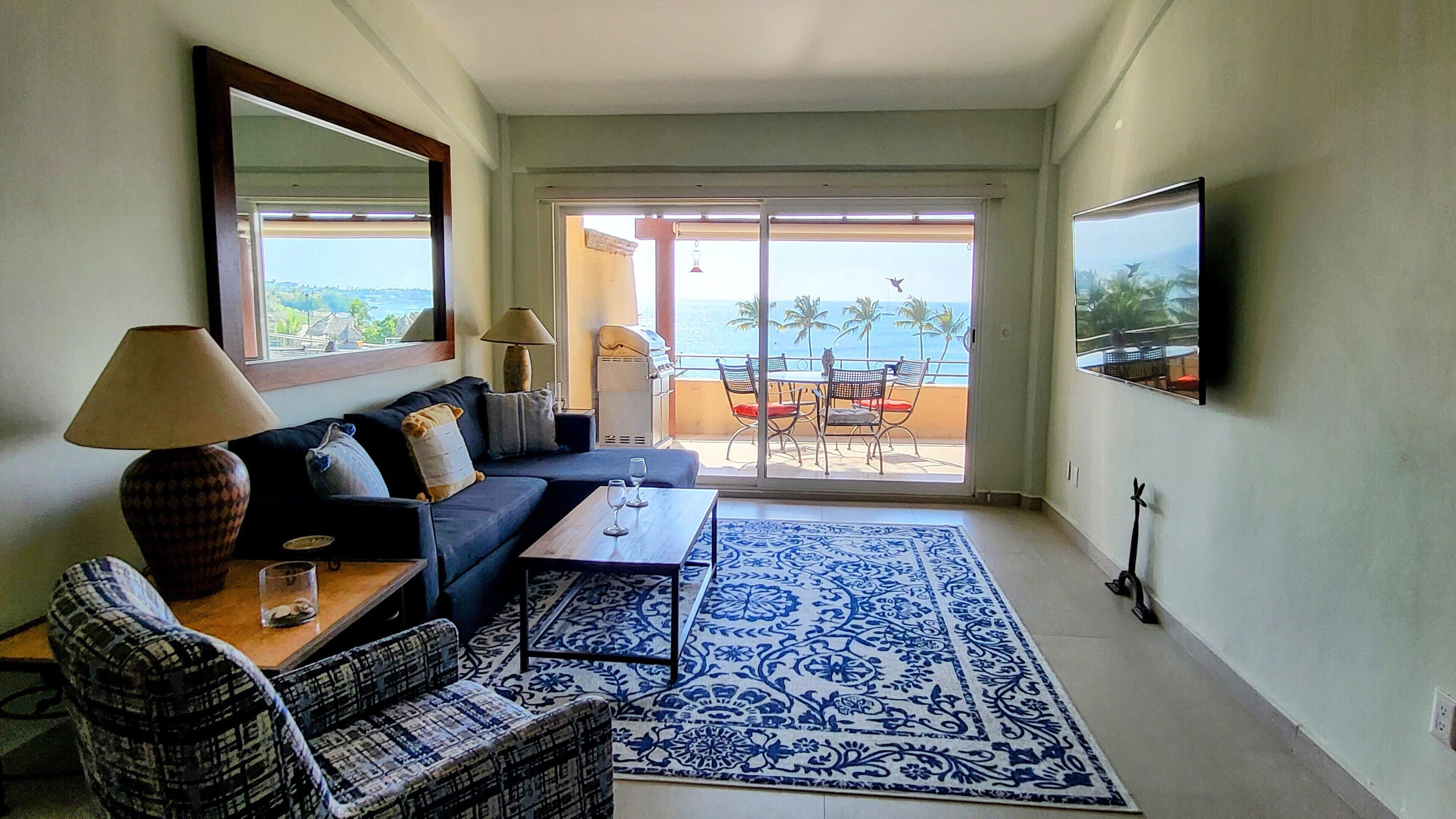 A living room with a sofa, coffee table, and patterned rug. There is a balcony with a view of the ocean and palm trees.