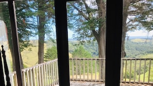 A view from a room with a large window overlooking a wooden deck with a white railing, surrounded by trees and a hilly landscape. - Large, genuine, Log House. 857 Church Rd. Kaitaia (Kaitaia)