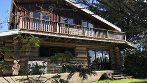 A log cabin with a balcony, stone foundation, and surrounding greenery. - Large, genuine, Log House. 857 Church Rd. Kaitaia (Kaitaia)