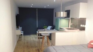 A modern, compact hotel room with a bed, a small kitchenette, a dining area, and a darkened living space.
