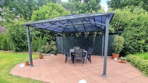 A covered patio area with a table and chairs, surrounded by potted plants and a fence.