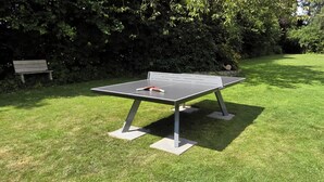 A ping pong table in a grassy outdoor area with a bench and dense foliage in the background.