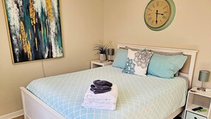 Una cama bien hecha con una colcha estampada, un reloj en la pared y una mesita de noche con una lámpara.