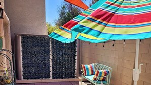 Un patio al aire libre con piso de ladrillo, una silla blanca con un cojín de colores, una pequeña mesa redonda y un paraguas a rayas que da sombra.