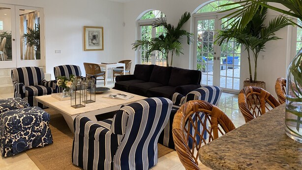Sala de estar com sofás listrados, um sofá preto, uma mesa de centro e uma área de jantar com uma mesa e cadeiras brancas. Há plantas em vasos e uma grande janela que proporciona luz natural.