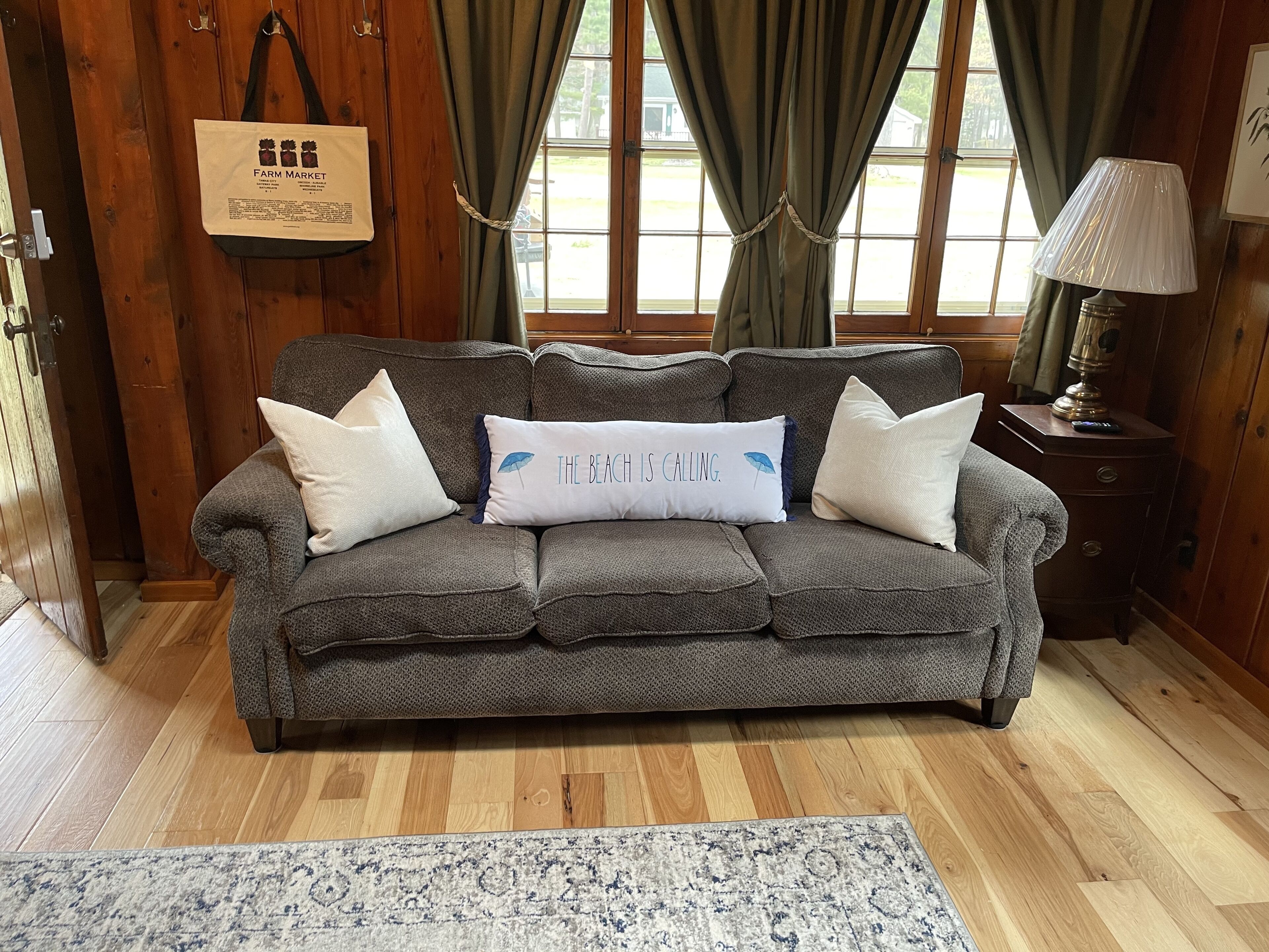 A grey sofa with a pillow that reads 'THE BEACH IS CALLING.' in front of a window with curtains.