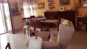 A living room with a dining area, a sofa, a coffee table, and a fireplace. There are framed pictures on the wall and a window with curtains. - House (Agropoli)