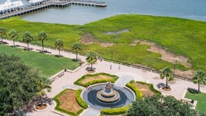 Un parque frente al agua con una fuente circular, un muelle y un barco grande al fondo.