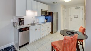 Una cocina compacta con armarios blancos, microondas, refrigerador, lavavajillas y una zona de comedor con dos sillas naranjas y una mesa.