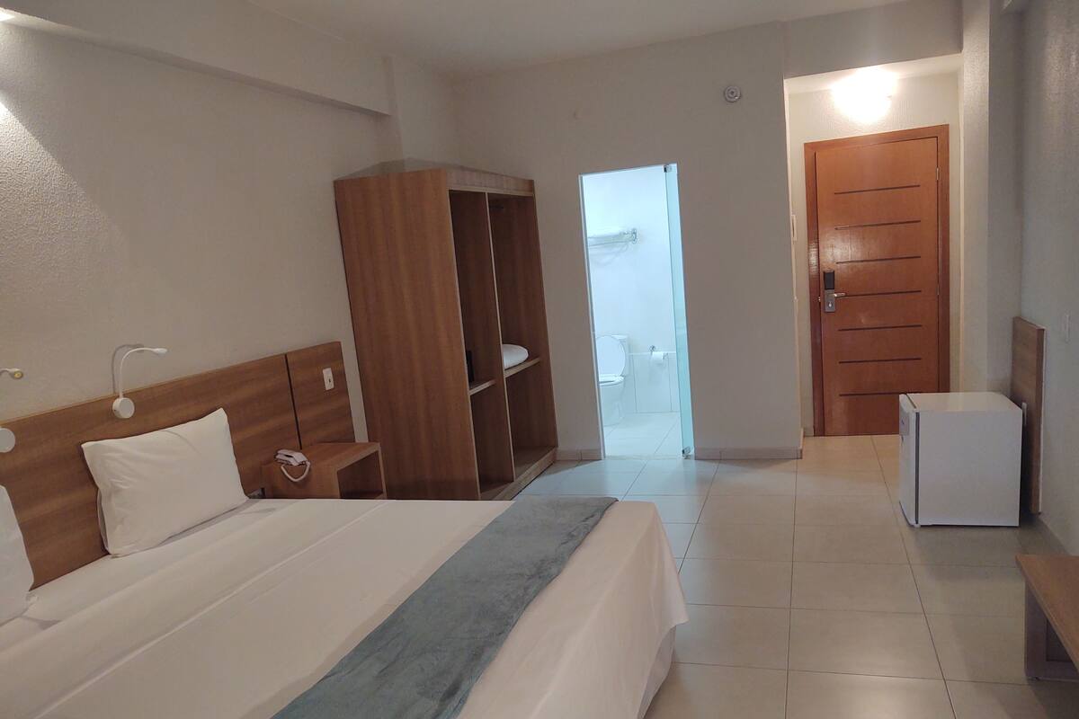 Une chambre d’hôtel avec un lit, une armoire en bois, un petit réfrigérateur blanc et une salle de bain visible à travers une porte ouverte.