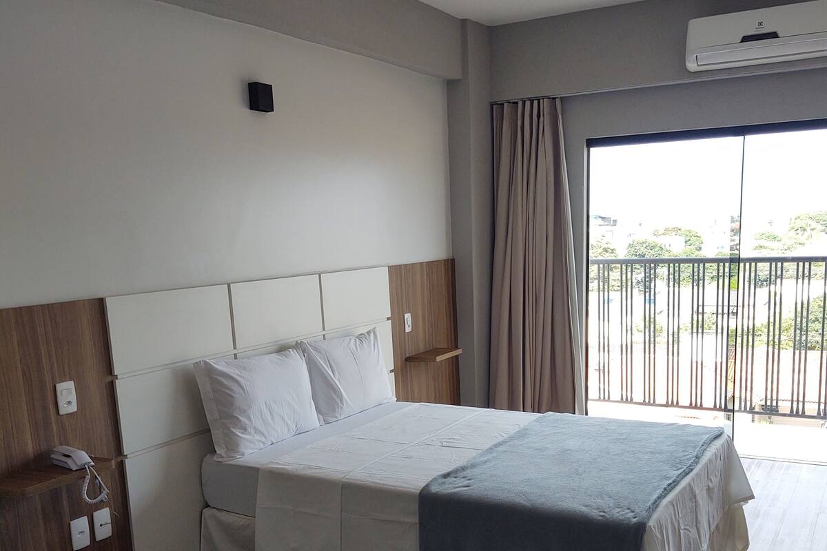 Une chambre d’hôtel équipée d’un lit, d’un téléphone, d’un climatiseur mural et offrant une vue sur l’extérieur par une fenêtre avec des rideaux.