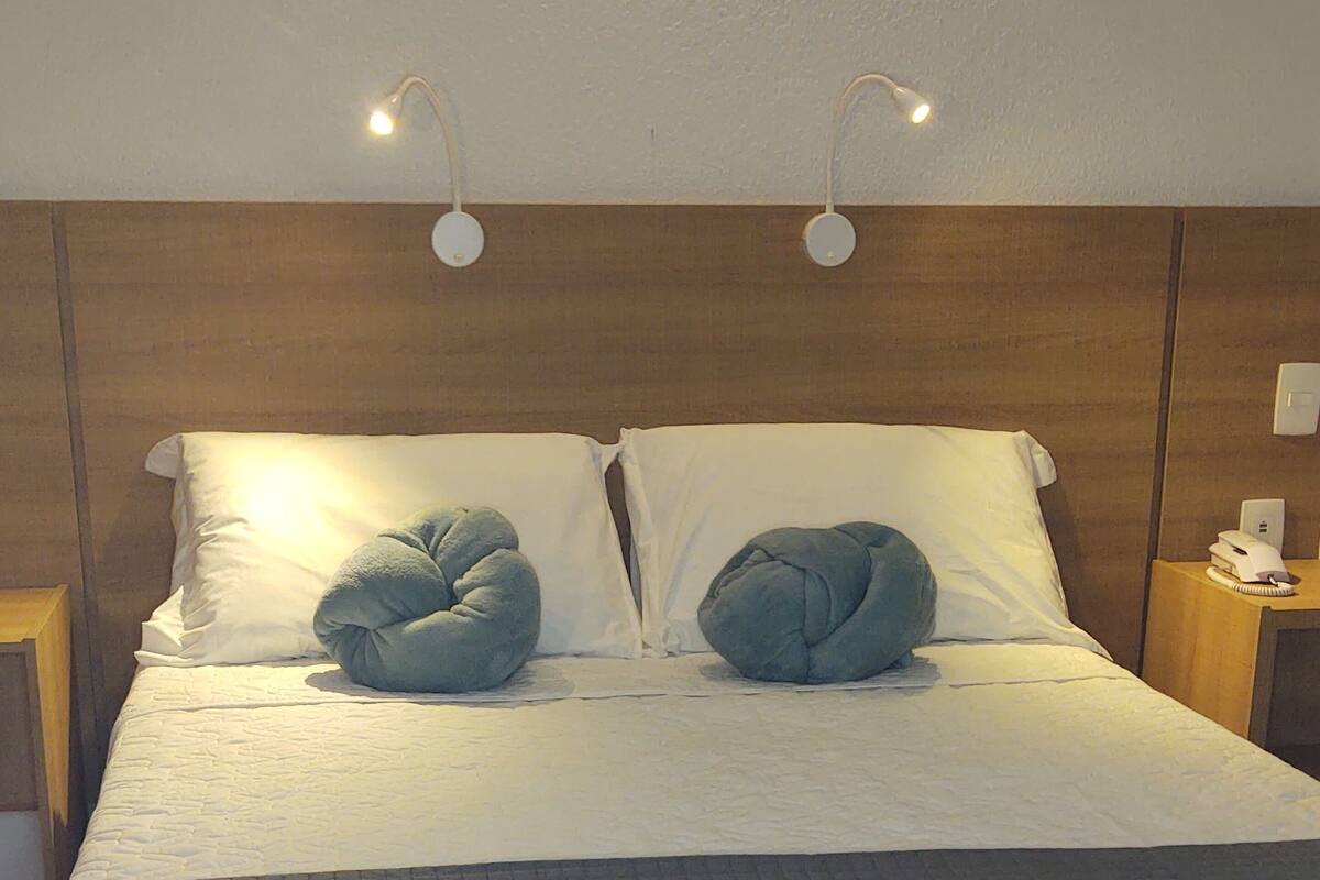Un lit bien fait, avec une tête de lit matelassée, deux oreillers et une table de chevet surmontée d’une lampe.