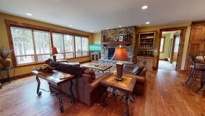 A spacious living room with a stone fireplace, wooden furniture, and large windows offering a view of the outdoors. - Cozy Family Escape with Access to Exclusive Golf (Sunriver)