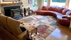 A cozy living room with a fireplace, leather sofa, and a large window with a view of greenery. - Mt. Alice Room at Bell In The Woods B&B (Queen / Second Floor )Max 2ppl (Seward)