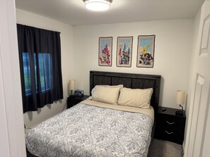 A bedroom with a bed, a nightstand, a lamp, a window with curtains, and three framed floral paintings on the wall.