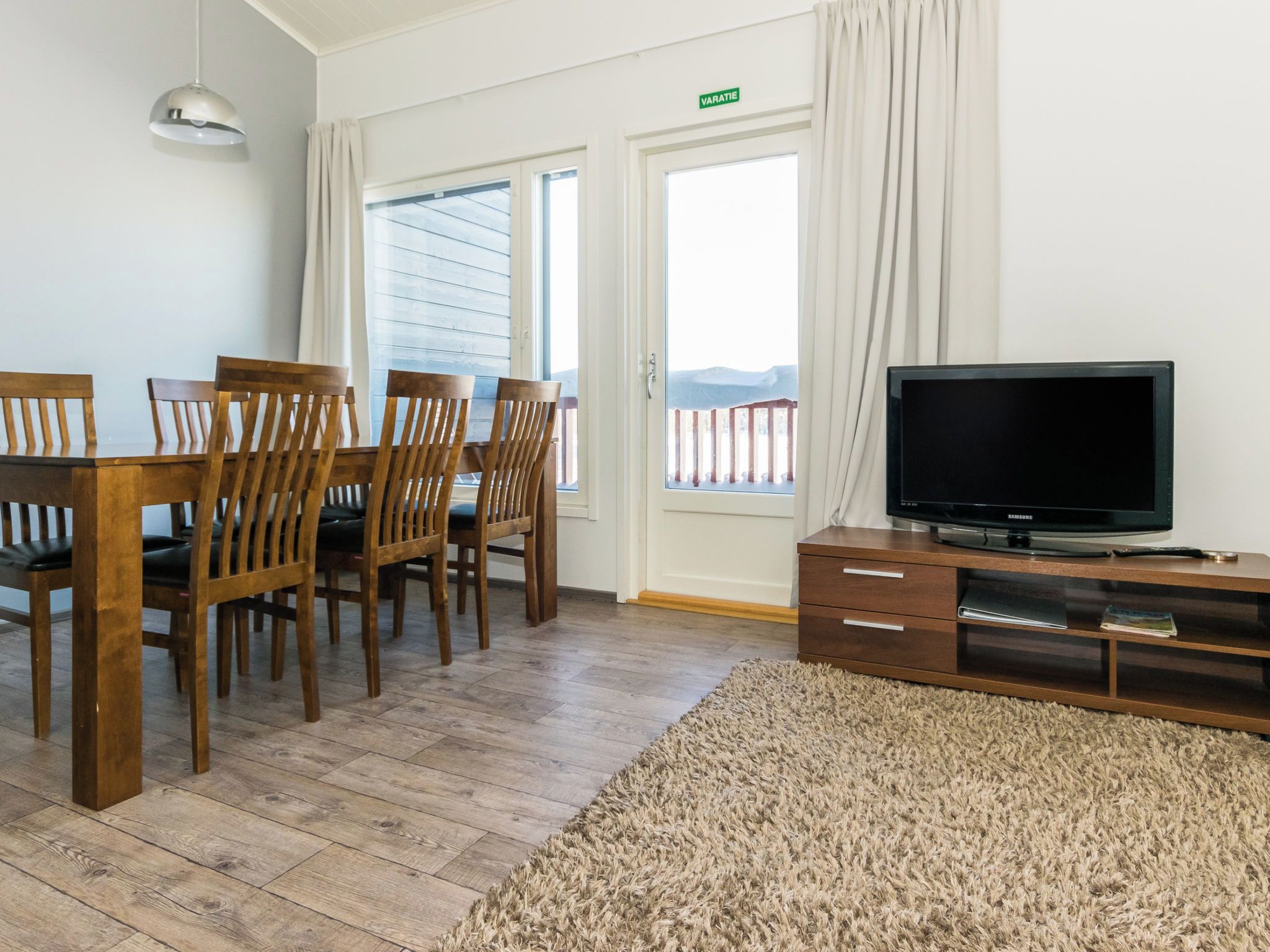 Een kamer met een houten eettafel en stoelen, een flatscreen televisie op een houten dressoir, en een groot raam met gordijnen.