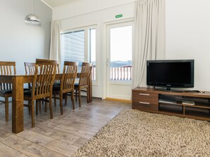 A room with a wooden dining table and chairs, a flat-screen TV on a wooden stand, and a large window with curtains.