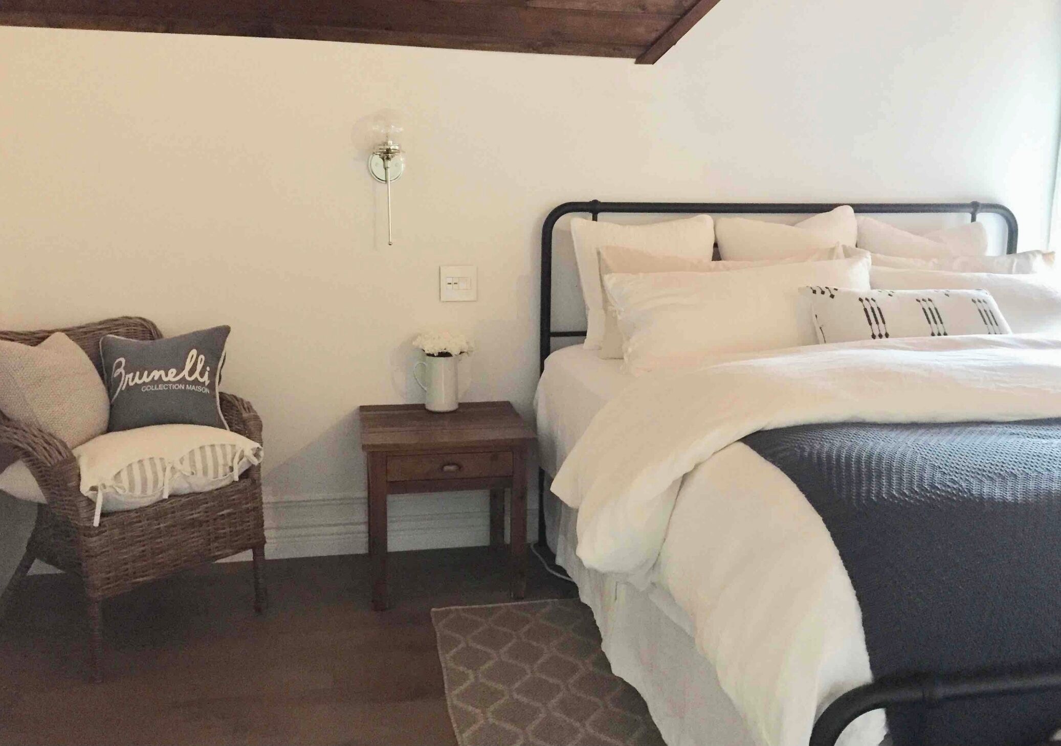 A bedroom with a bed, a nightstand, a wicker chair, and a wall-mounted light fixture.