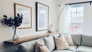 A modern living room with a grey sectional sofa, a wooden shelf with decorative items, and a large window with a view of a red structure outside.