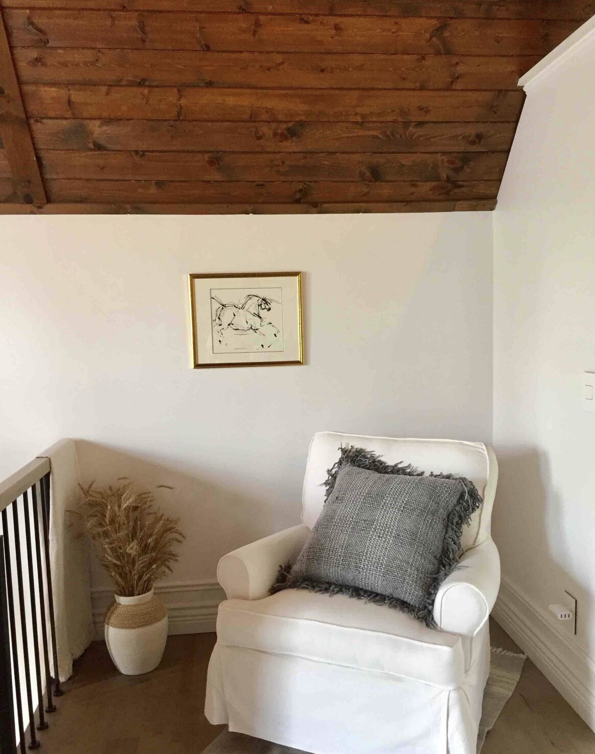 Un sillón blanco con un cojín gris, un boceto enmarcado en la pared y una jarra alta con plantas secas.