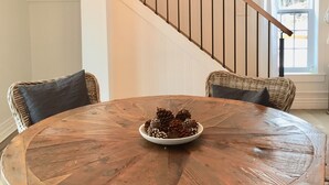 A round wooden dining table with a bowl of pine cones, surrounded by wicker chairs with cushions. A chandelier with four candles hangs above.