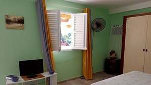 A bedroom with a bed, a TV on a stand, a fan, and a window with curtains. - Studio Paradis Caché Sainte-Marie Kybo Karaib Location pkg Mtgne (Sainte-Marie)