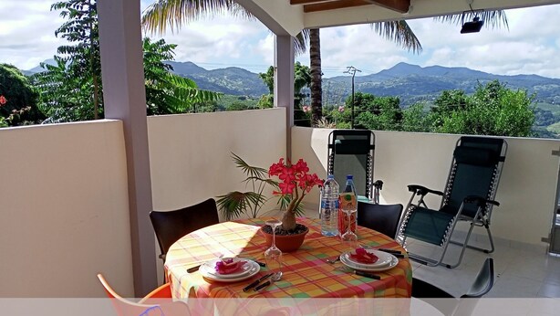 A balcony with a table set for two, a potted plant, and a view of mountains. - Studio Paradis Caché Sainte-Marie Kybo Karaib Location pkg Mtgne (Sainte-Marie)