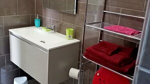 A modern bathroom with a white sink, a mirror, a toilet with a red sign, and a metal shelf with towels. - Studio Paradis Caché Sainte-Marie Kybo Karaib Location pkg Mtgne (Sainte-Marie)