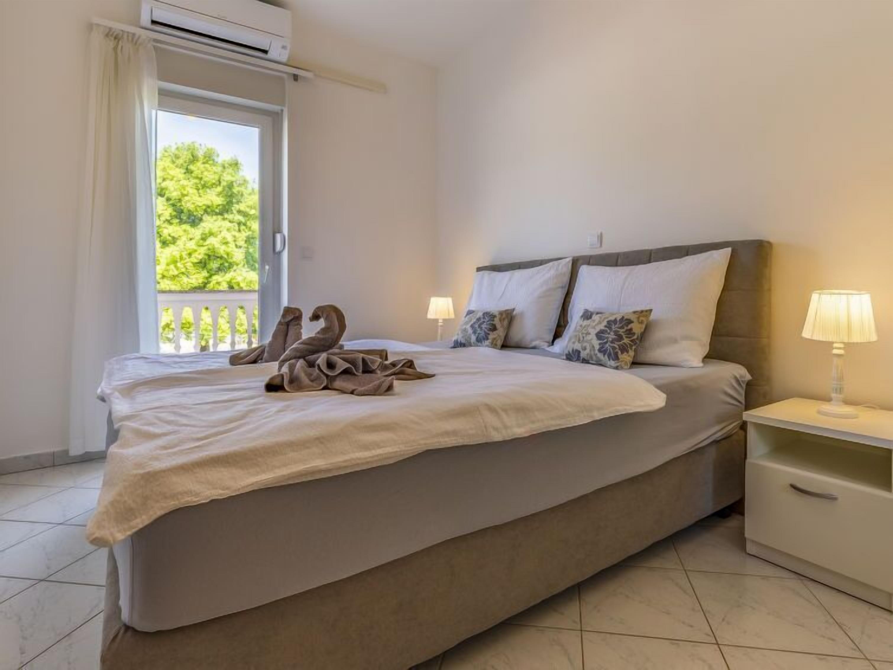 A neatly made bed with a blanket and towels, a bedside lamp, and a view of greenery outside.
