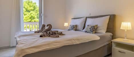 A neatly made bed with a blanket and towels, a bedside lamp, and a view of greenery outside.
