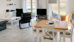 Un salon moderne avec une table à manger en bois, des chaises blanches, un canapé noir, un téléviseur et une grande fenêtre donnant sur l’extérieur.